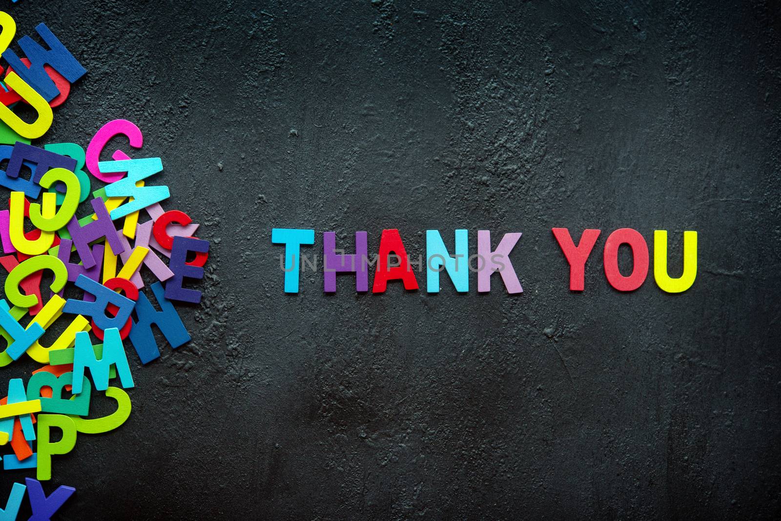 The colorful words "THANK YOU" made with wooden letters next to a pile of other letters over dark background.