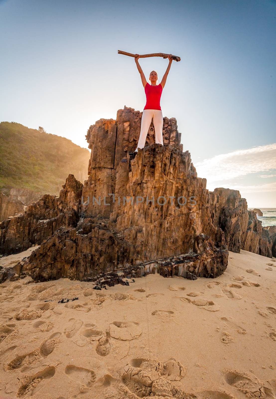 Energetic woman fitness health in beach location.  Standing on craggy rock with backlight of morning sun