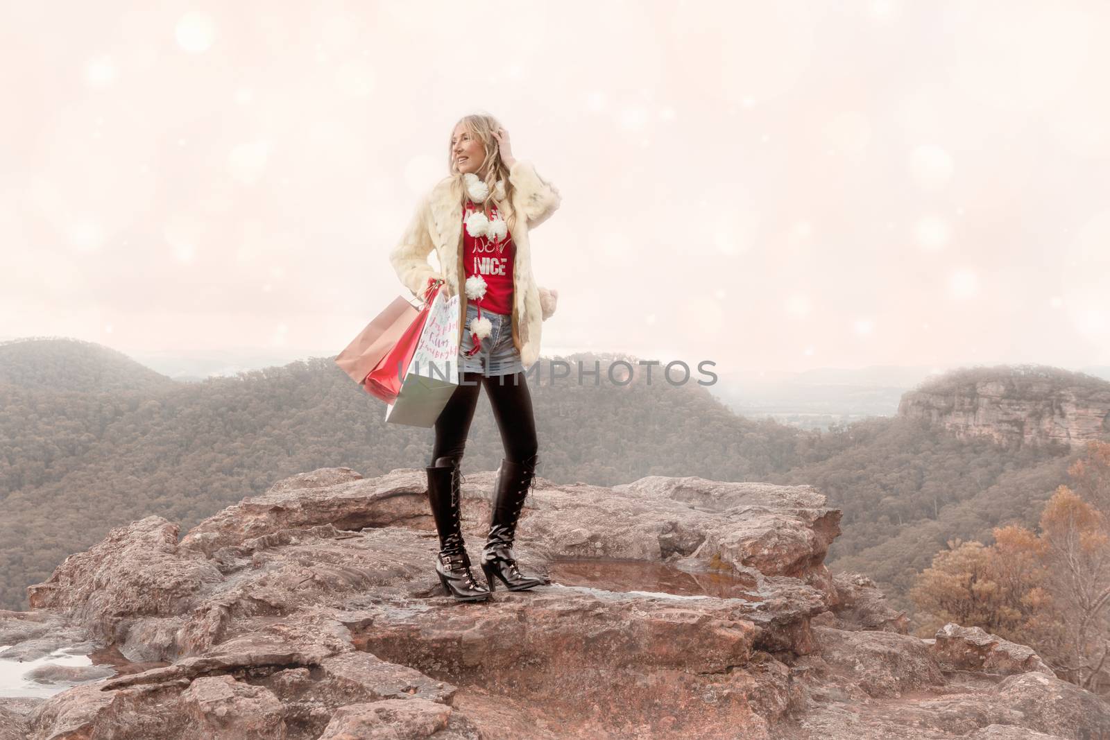 A magical Christmas in Blue Mountains. A female holding gift bags wearing warm coat, long boots denim shorts and a t-shirt with a wintry backdrop of mountains with soft bokeh. Space for copyChristmas themesBlue Mountains, winter magic, Christmas in July
