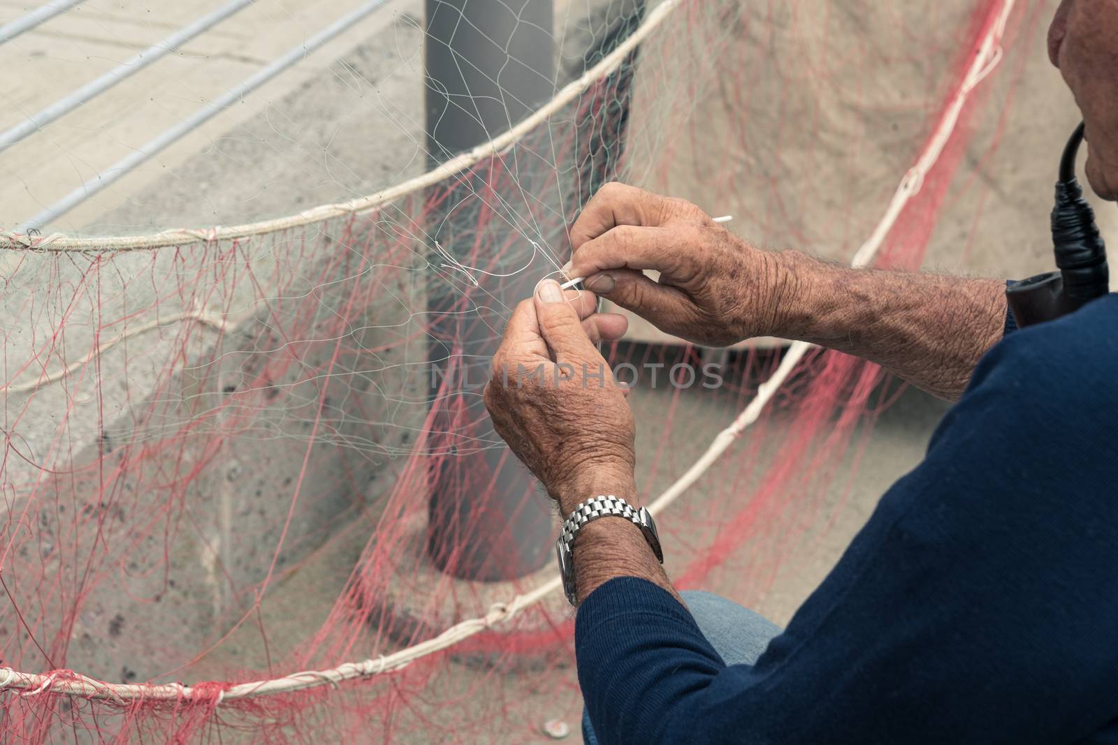 Old fisherman reparing fishing net, sitting with pipe in his mouth.