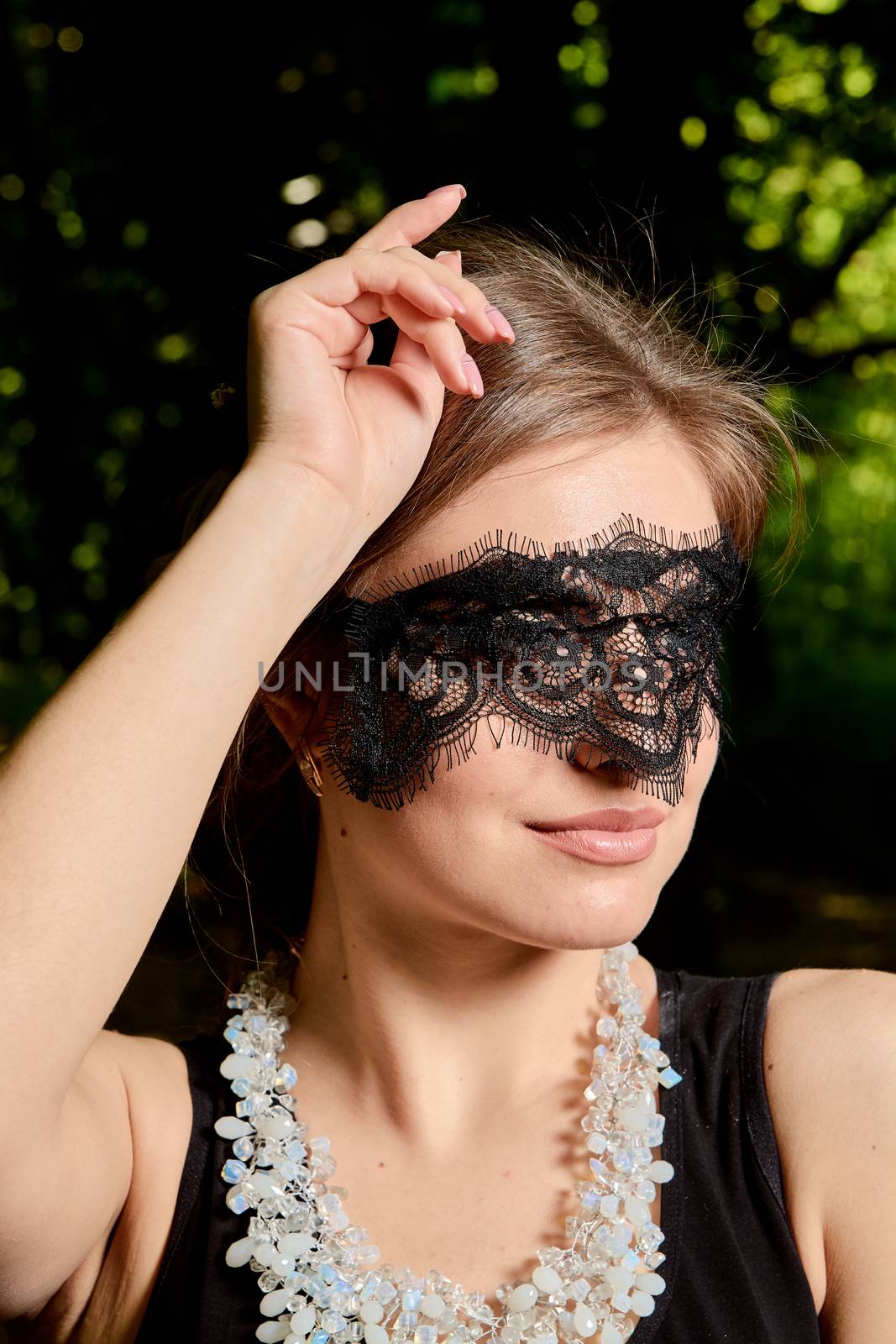 Young attractive woman in the sexy see-through, transparent, transpicuous, diaphanous, clear black dress and black lace masquerade venetian face mask posing outdoor at the green alley in the old park.
