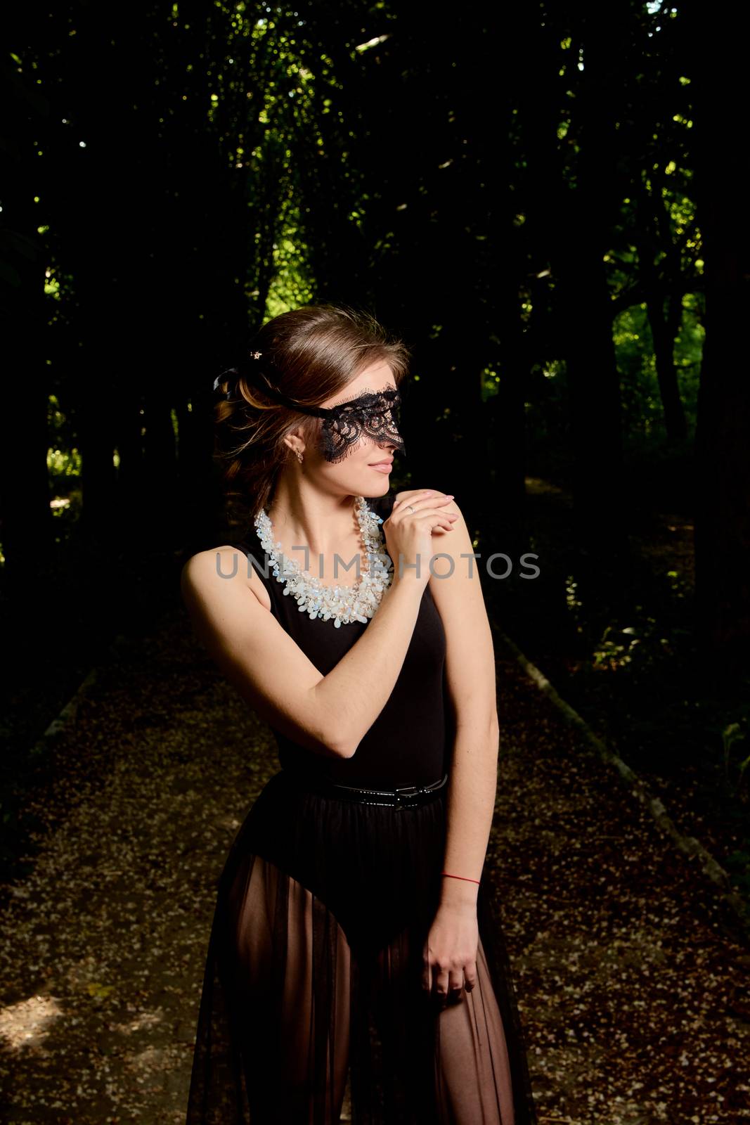 Young attractive woman in the sexy see-through, transparent, transpicuous, diaphanous, clear black dress and black lace masquerade venetian face mask posing outdoor at the green alley in the old park.