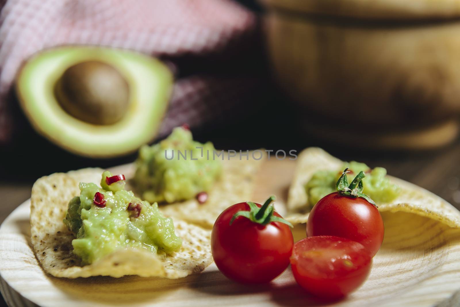 homemade nachos with guacamole in a wood plate to an unfocused avocado and a kitchen rag, typical mexican healthy vegan cuisine with rustic dark food photo style, selective focus