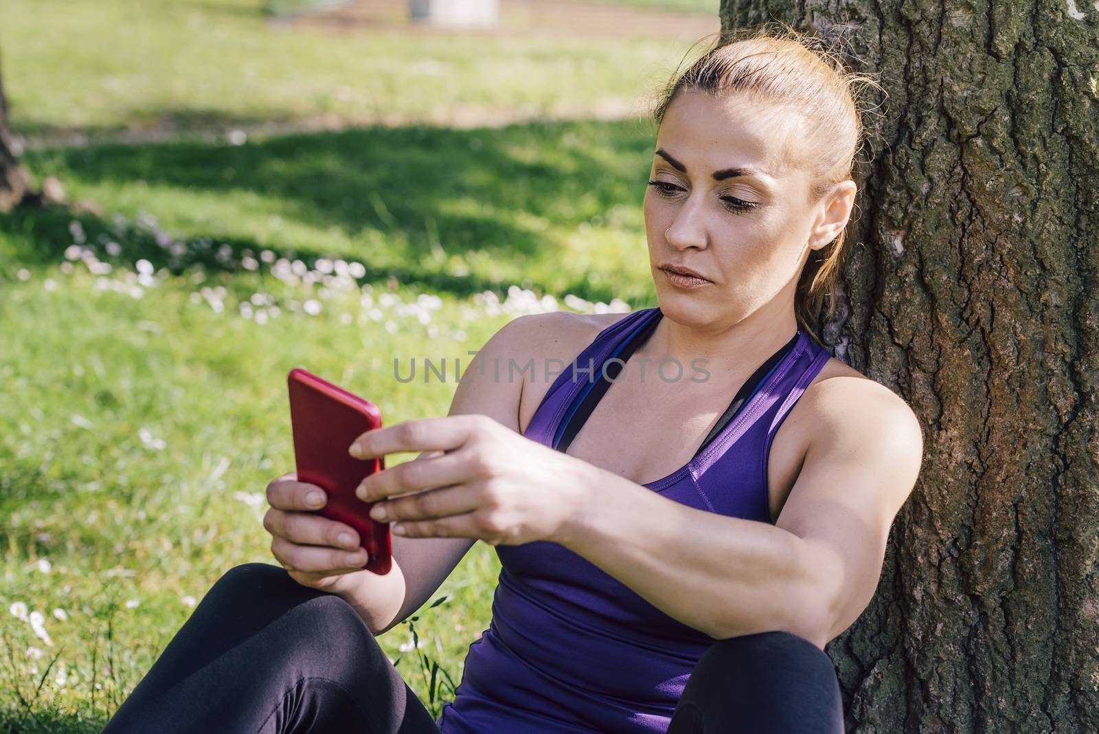 fitness woman taking a workout rest sitting next to a tree for texting on her phone, healthy modern lifestyle and sport concept