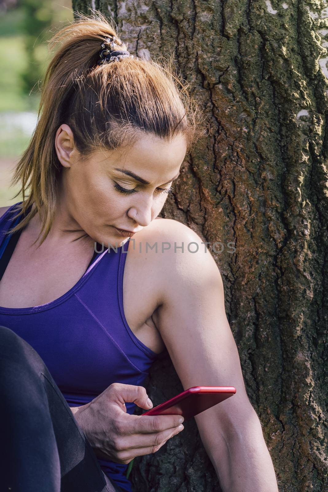 fitness woman taking a workout rest sitting next to a tree for take a look to her phone, healthy modern lifestyle and sport concept