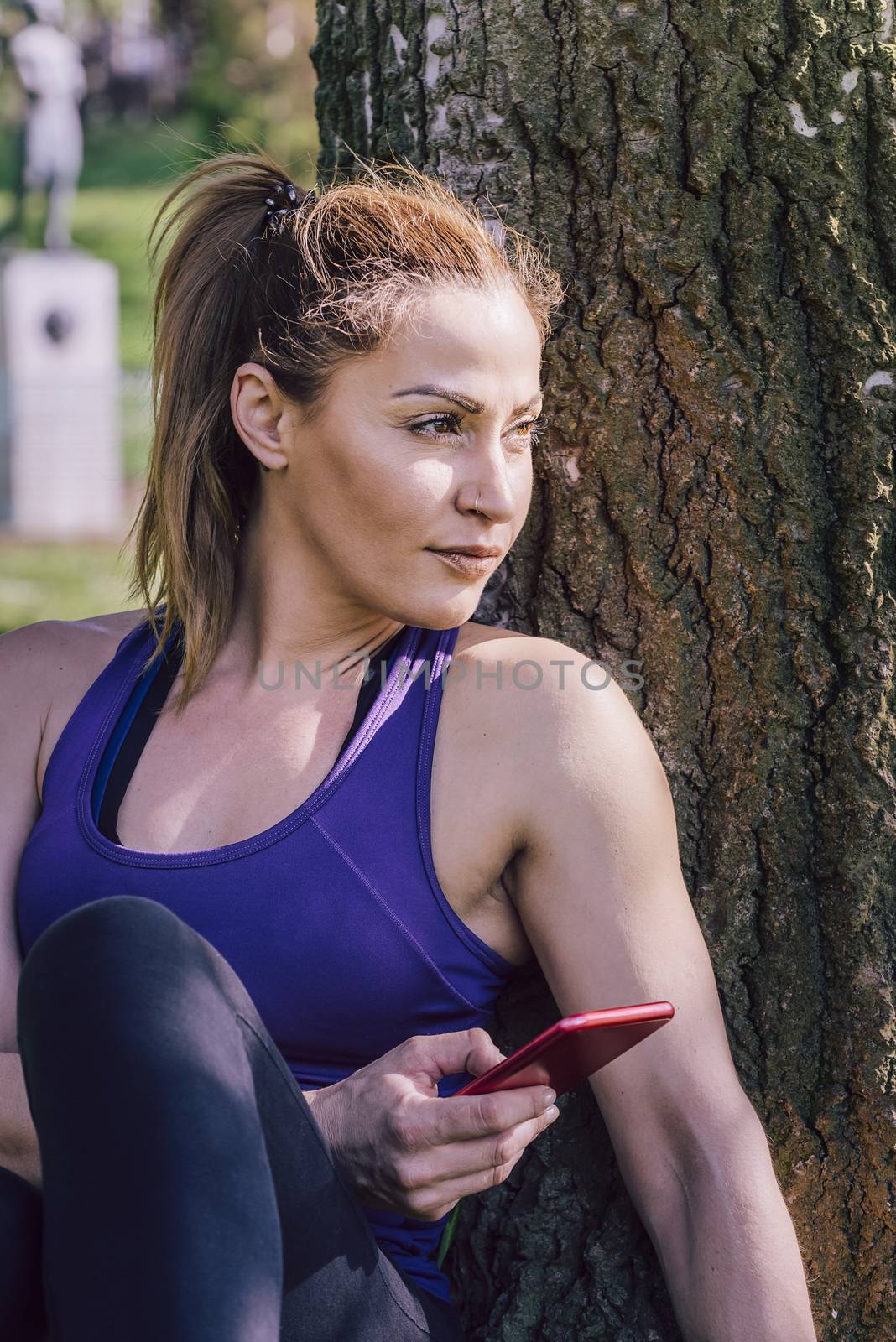 sports woman resting after exercise to take a look to her phone while sitting next to a tree, healthy modern lifestyle and sport concept