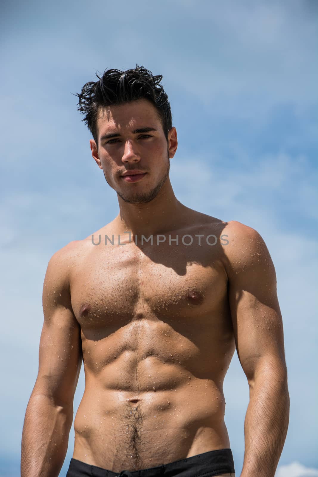 Young fit athletic man at beach in summer day showing muscular torso, looking at camera