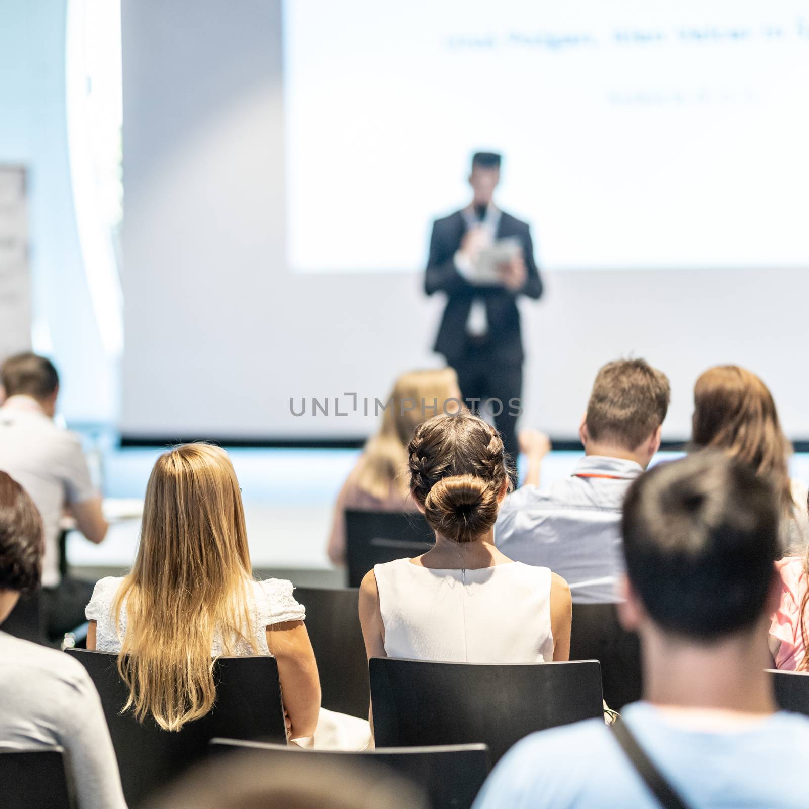 Business and entrepreneurship symposium. Speaker giving a talk at business meeting. Audience in conference hall. Rear view of unrecognized participant in audience.