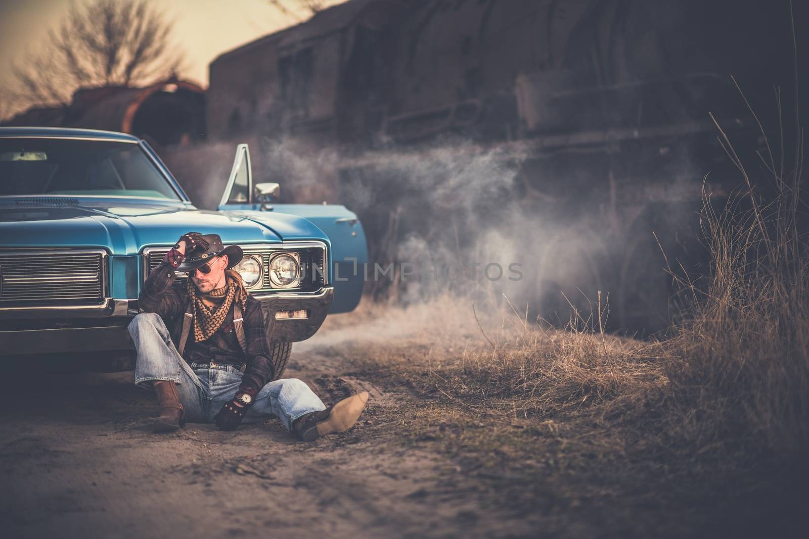 The American Cowboy Story. Caucasian Men Wearing Western Style Clothes and Cowboy Hat, Taking a Moment in Front of His American Classic Muscle Car. Aged and Rusty Steam Locomotive in the Background.