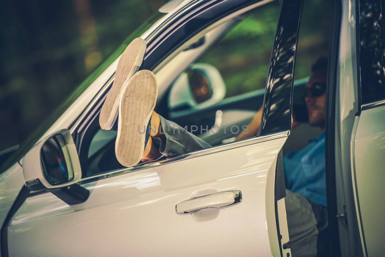 Leaving Stress Free Life. Caucasian Men Enjoying His Free Time in the Local Park While Laying Behind the Car Wheel with Legs Out of the Vehicle Window.