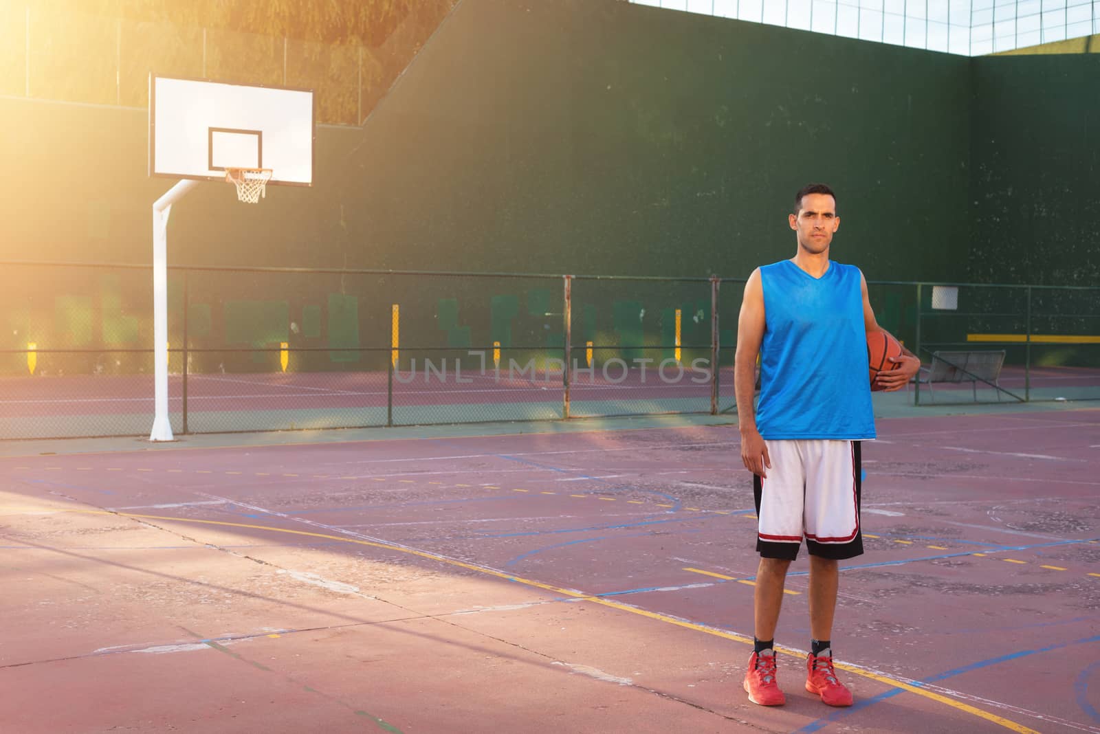 Athlete man holding basketball ball standing on playground at sunset by HERRAEZ