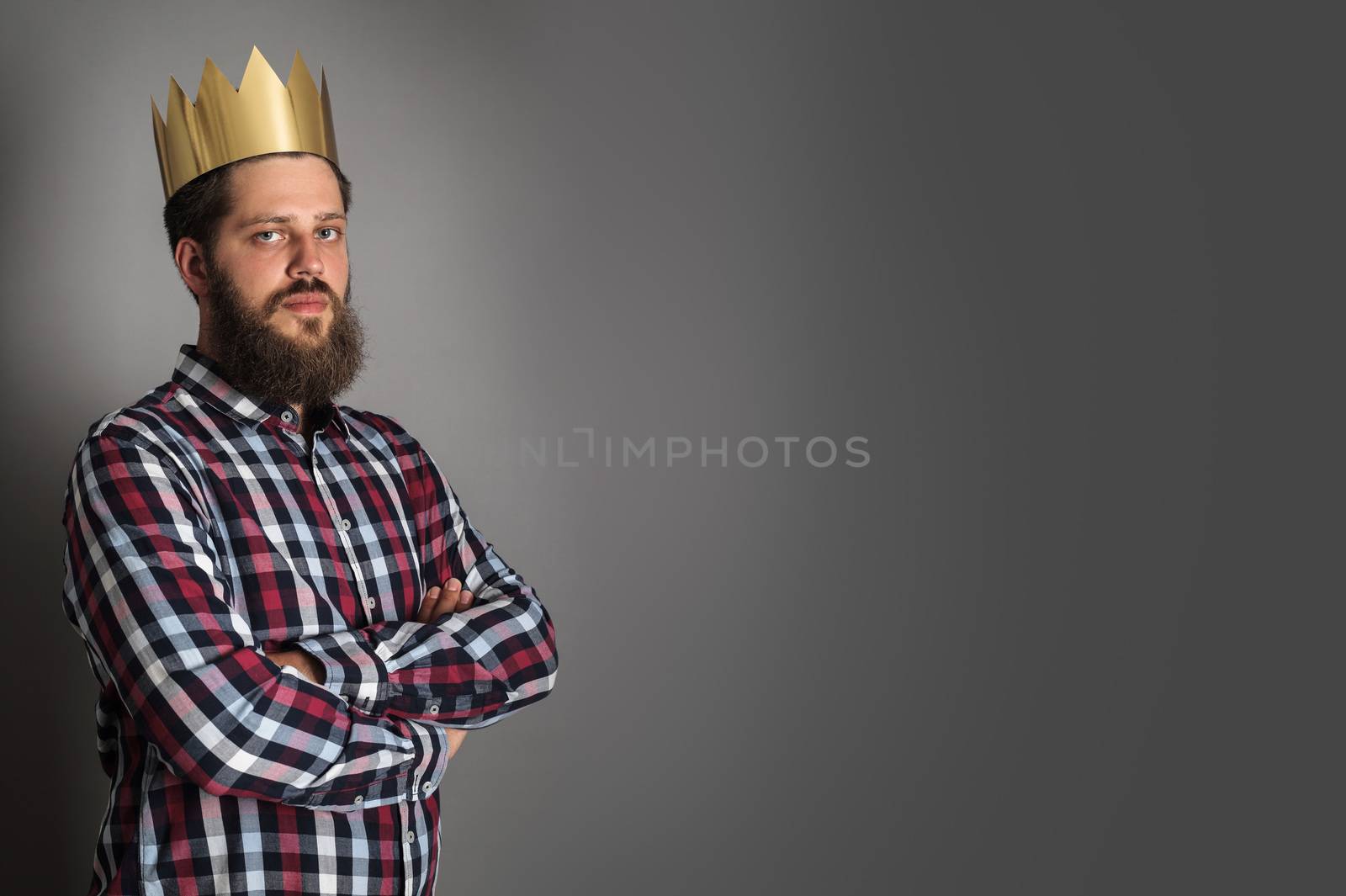 Bearded man in golden crown looking at camera while standing with crossed arms on gray background