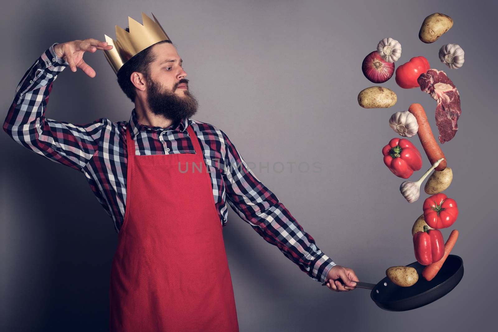 Cooking man concept, king of kitchen,bearded man in checked shirt, drop up meat and vegetables from a pan, studio shot on gray background