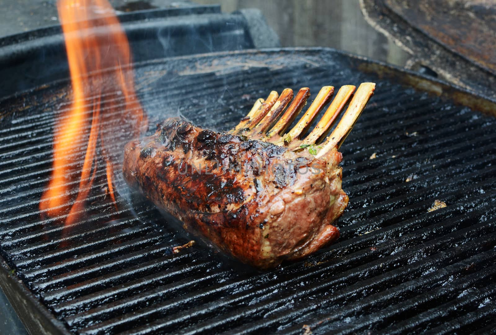 Flame rises from a grill around barbecued meat - a rack of lamb with 7 ribs