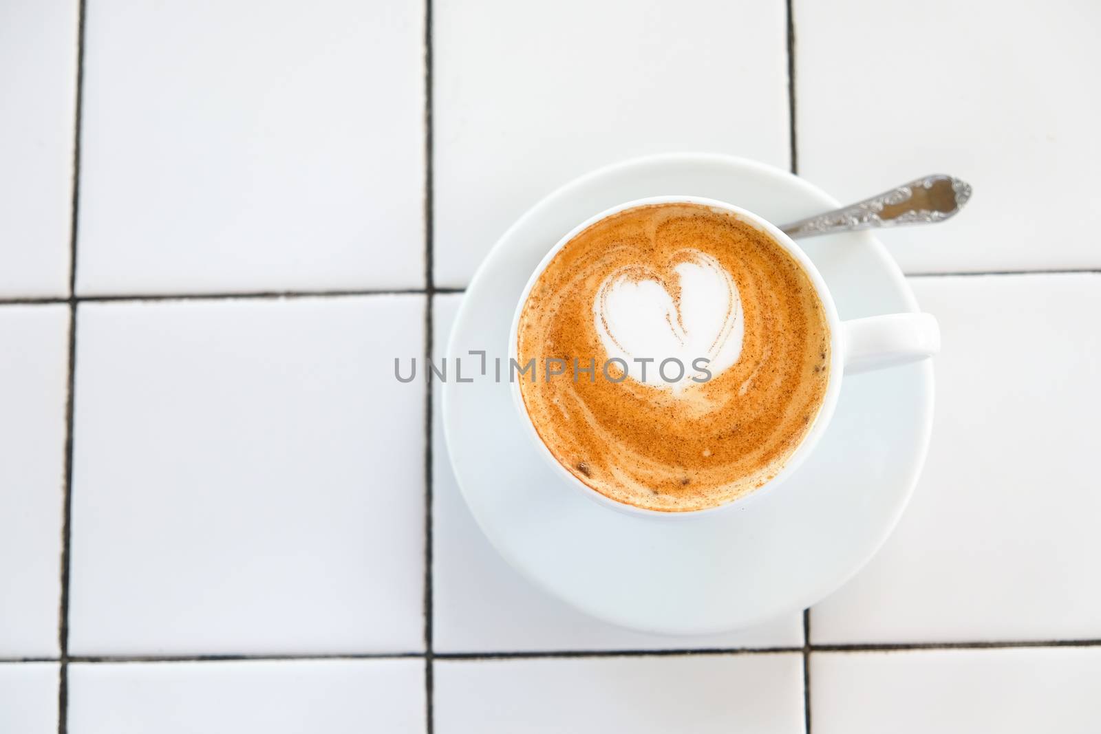 Cappuccino cup on tiled white table background. Foam is decorated with cinnamon heart. Copy space. Top view, located at side of frame. Horizontal. Mock up for social media, food blog, lifestyle.