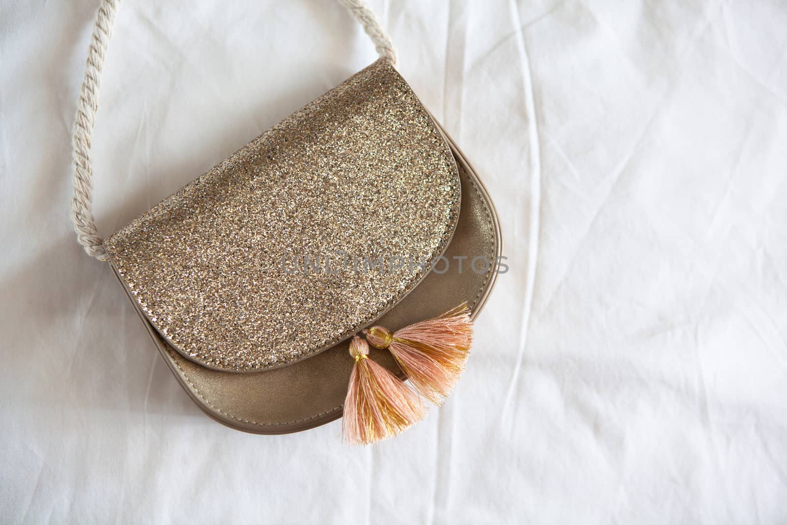 Small golden saddle handbag with tassels and rope handle lies on rumpled white sheet. Concept fees, outfitting, kid's fashion, basic wardrobe. Copy space. Flat lay, top view