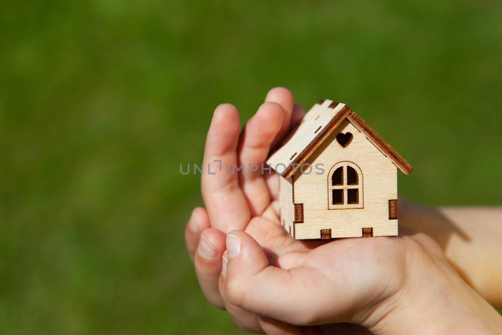 Small toy house in hands of child on green grass background. Concept mortgage, dream house, real estate acquisition. In frame only hands, without face . Soft selective focus. Close-up. Copy space.