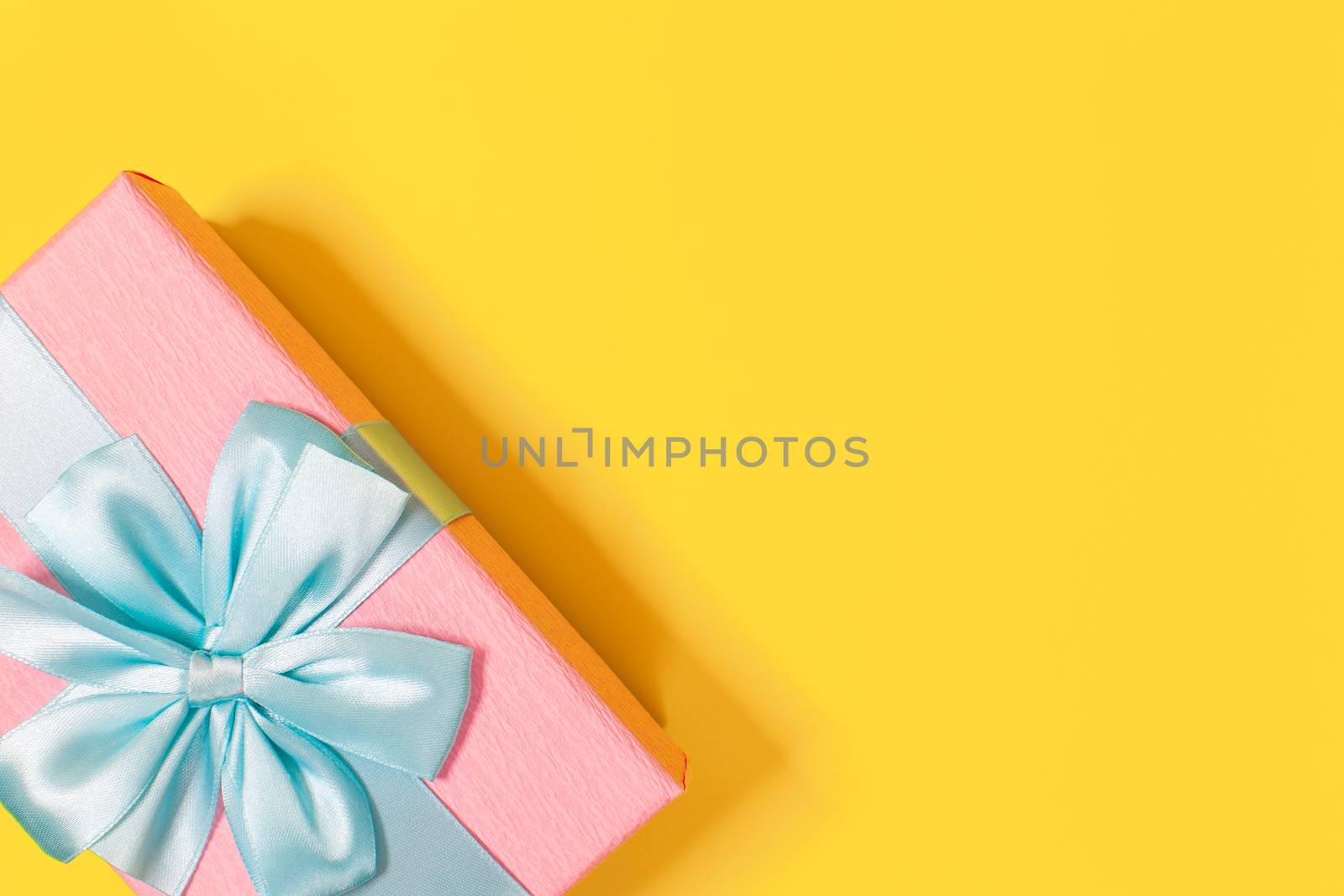 Pink gift box tied with blue ribbon with bow at the top on yellow background. Copy space for text. Minimal flat lay. Top view. Birthday, New year, Mother's day, Women's day celebration concept.