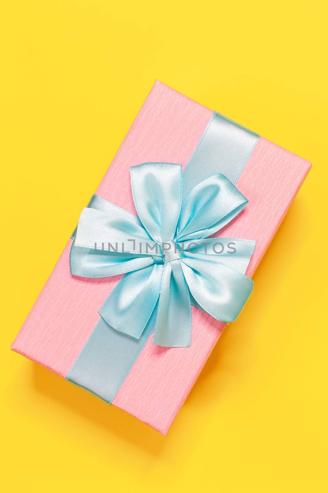 Pink gift box tied with blue ribbon with bow at the top on yellow background. Vertical. Minimal flat lay. Top view. Birthday, New year, Mother's day, Women's day celebration concept.