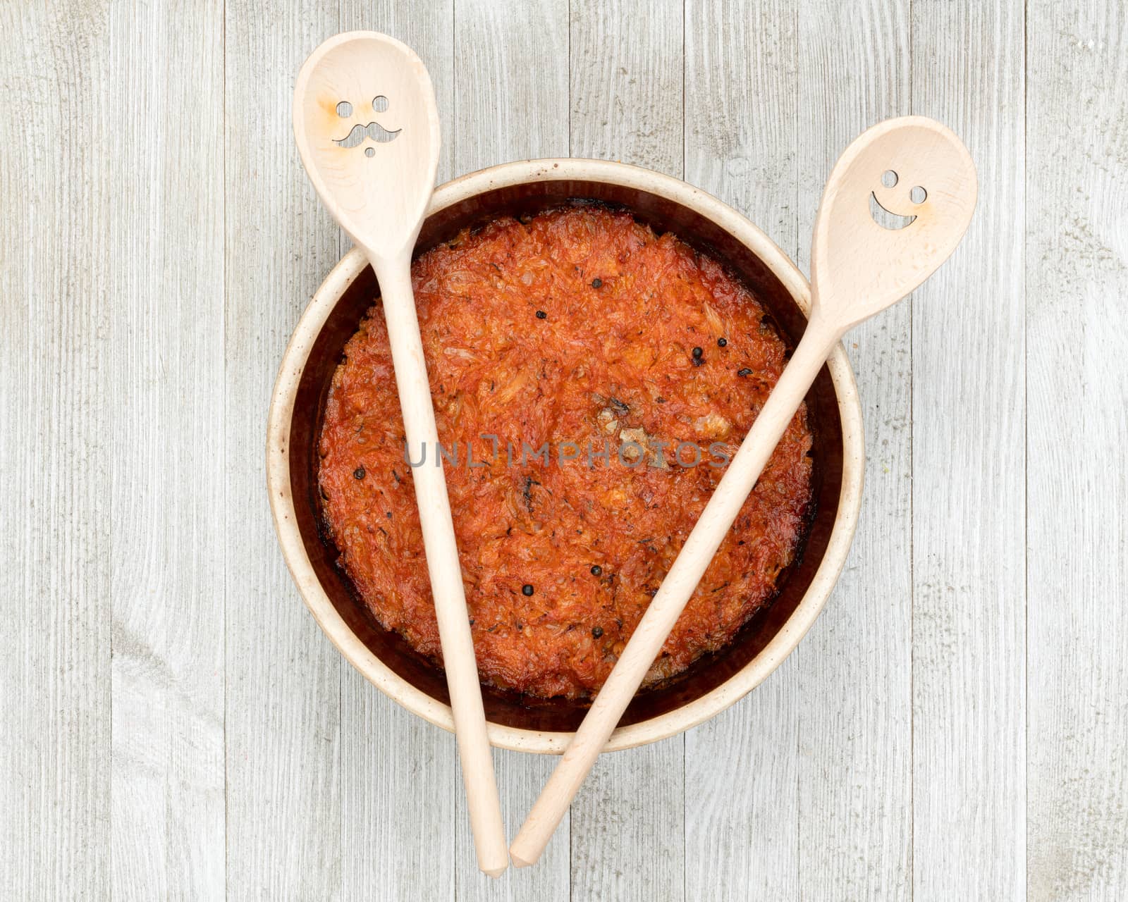 A ceramic pot of traditional Eastern European stewed cabbage and decorated wooden spoons on a white wooden kitchen board.