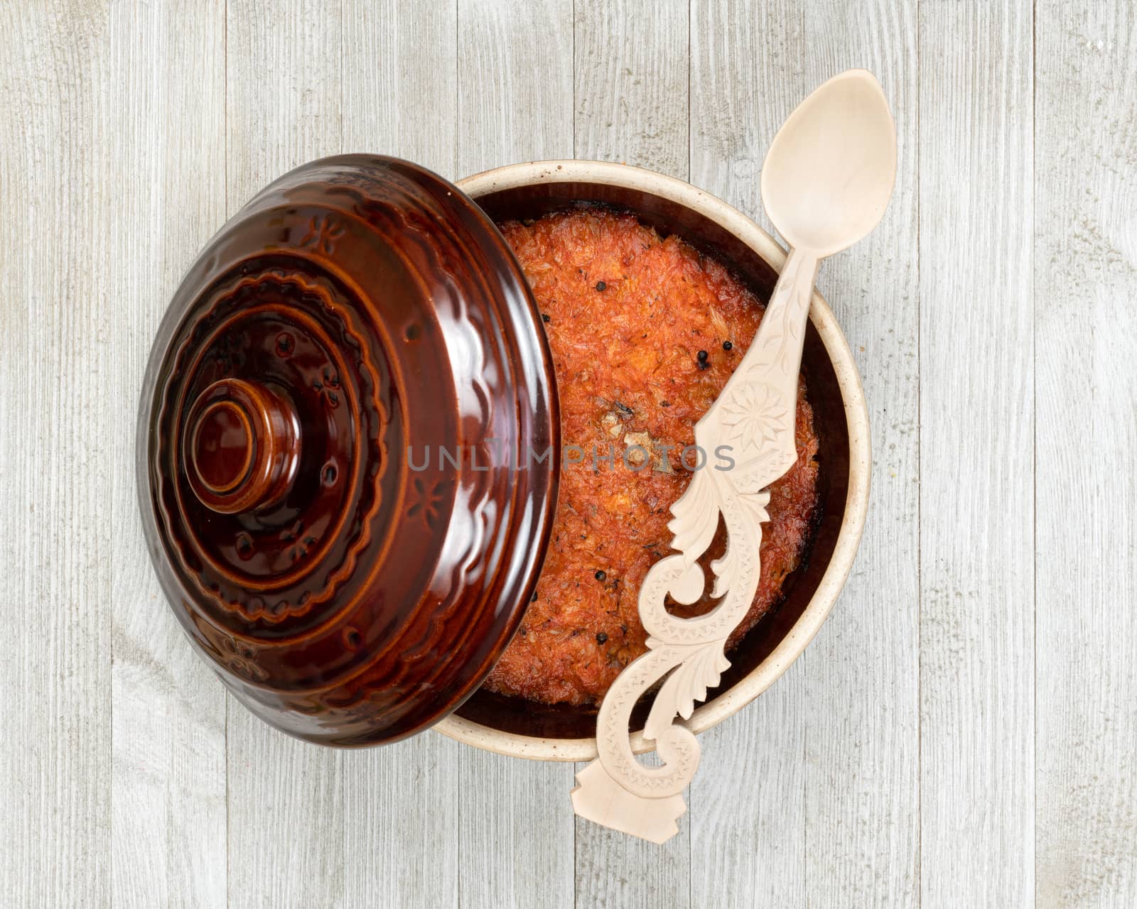 A ceramic pot of traditional Eastern European stewed cabbage and decorated wooden spoon on a white wooden kitchen board.