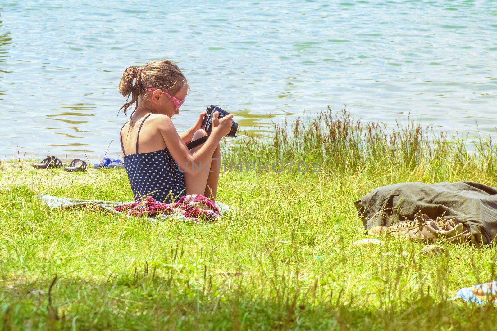 Little girl taking pictures sitting near the river on the grass.Cute little girl sitting on the grass on sunny summer day and taking picture with camera. Summer holidays.