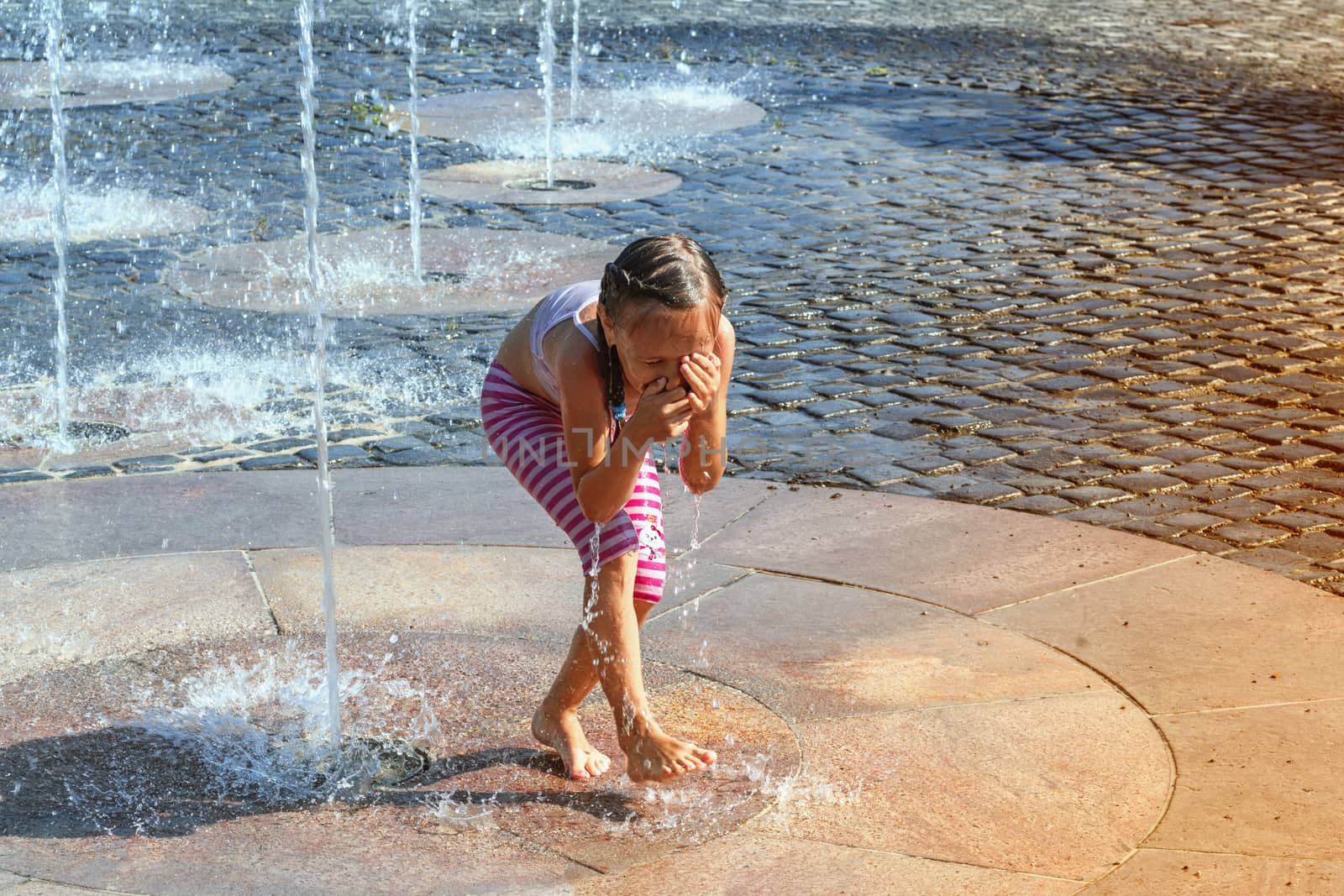 Girl on a sunny summer day are poured water from a fountain.Girl happily in shallow clean water on of city fountain on warm bright summer day.