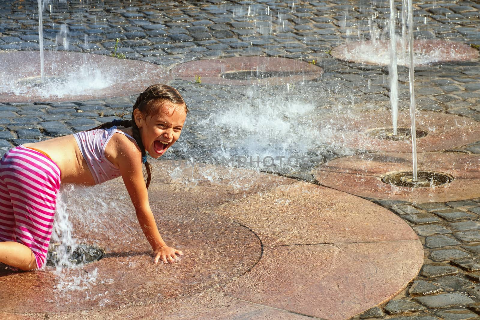 Girl on a sunny summer day are poured water from a fountain.Girl happily in shallow clean water on of city fountain on warm bright summer day.