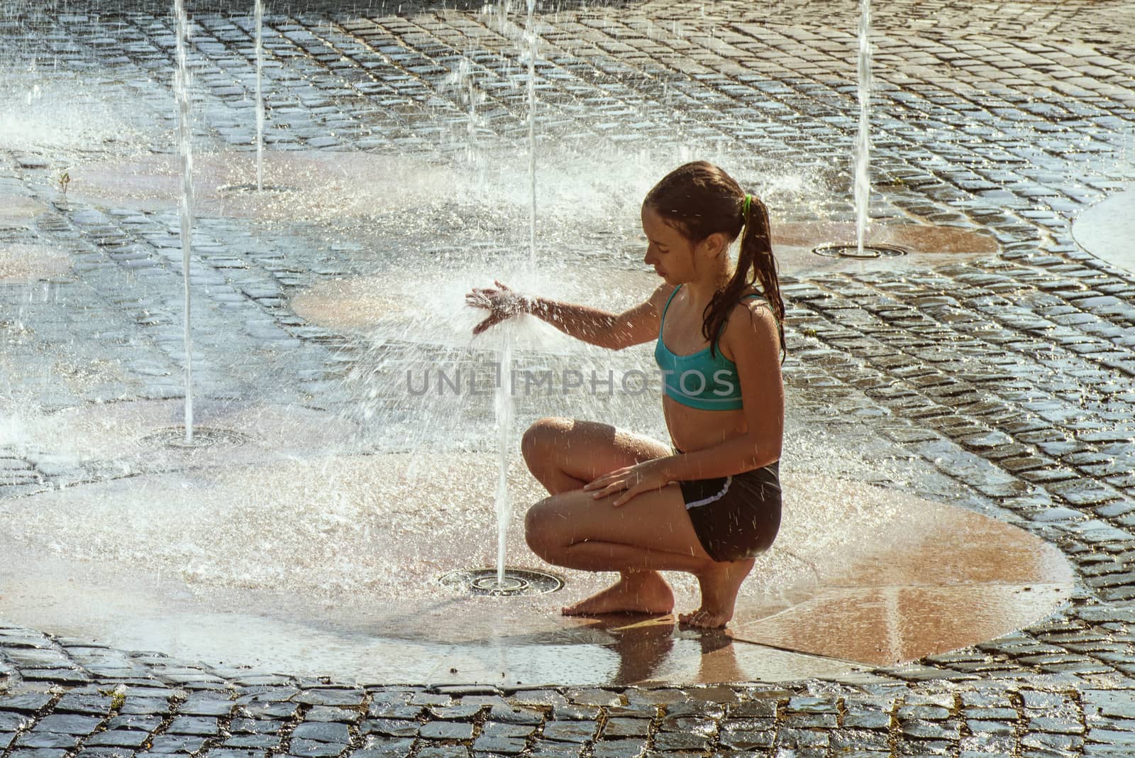Girl on a sunny summer day are poured water from a fountain.Girl happily in shallow clean water on of city fountain on warm bright summer day.