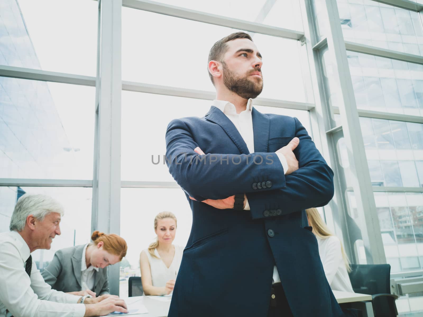 Confident young businessman standing with folded arms smiling , team of his colleagues sitting at the table in the background