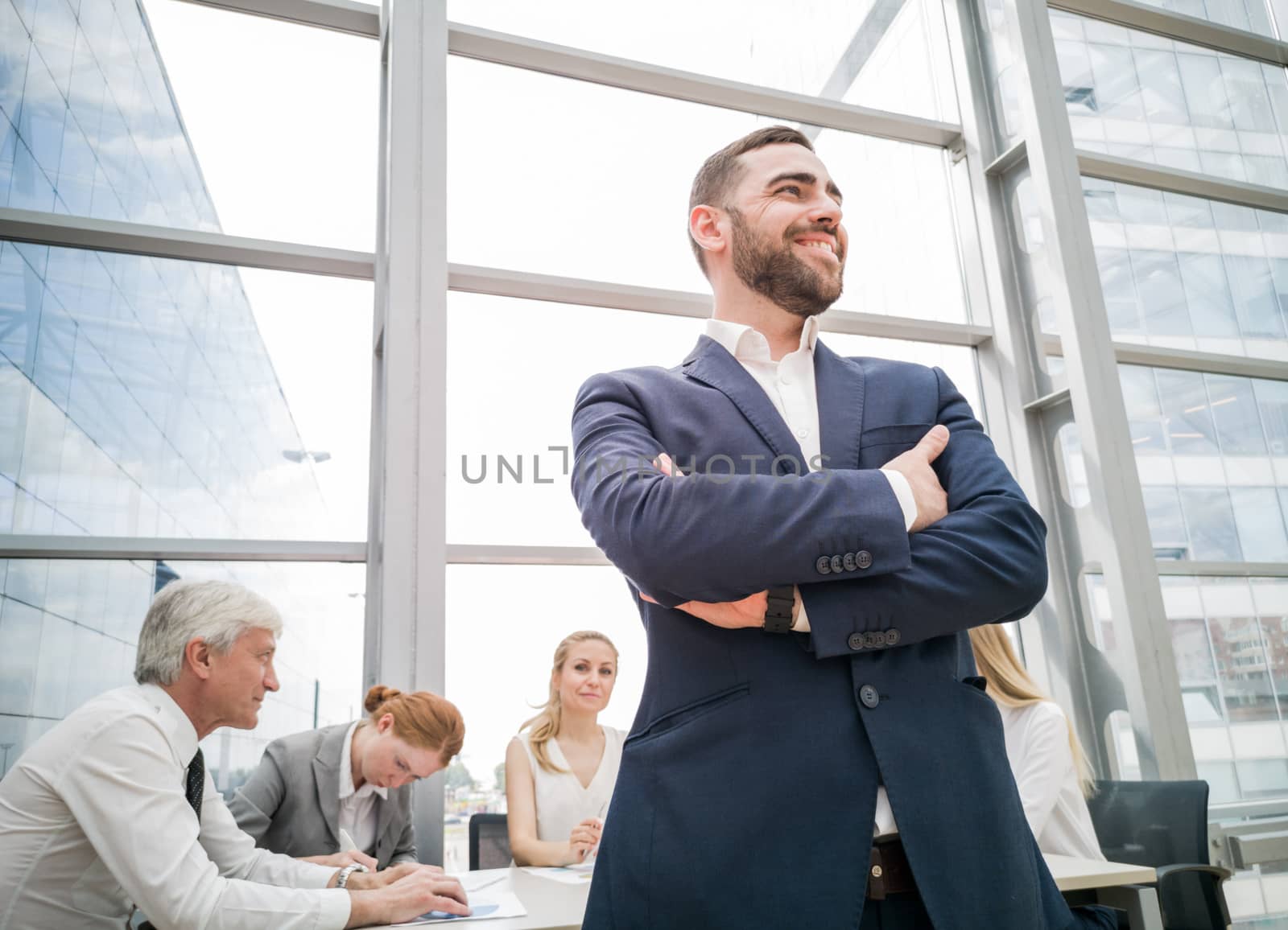 Confident young businessman standing with folded arms smiling , team of his colleagues sitting at the table in the background