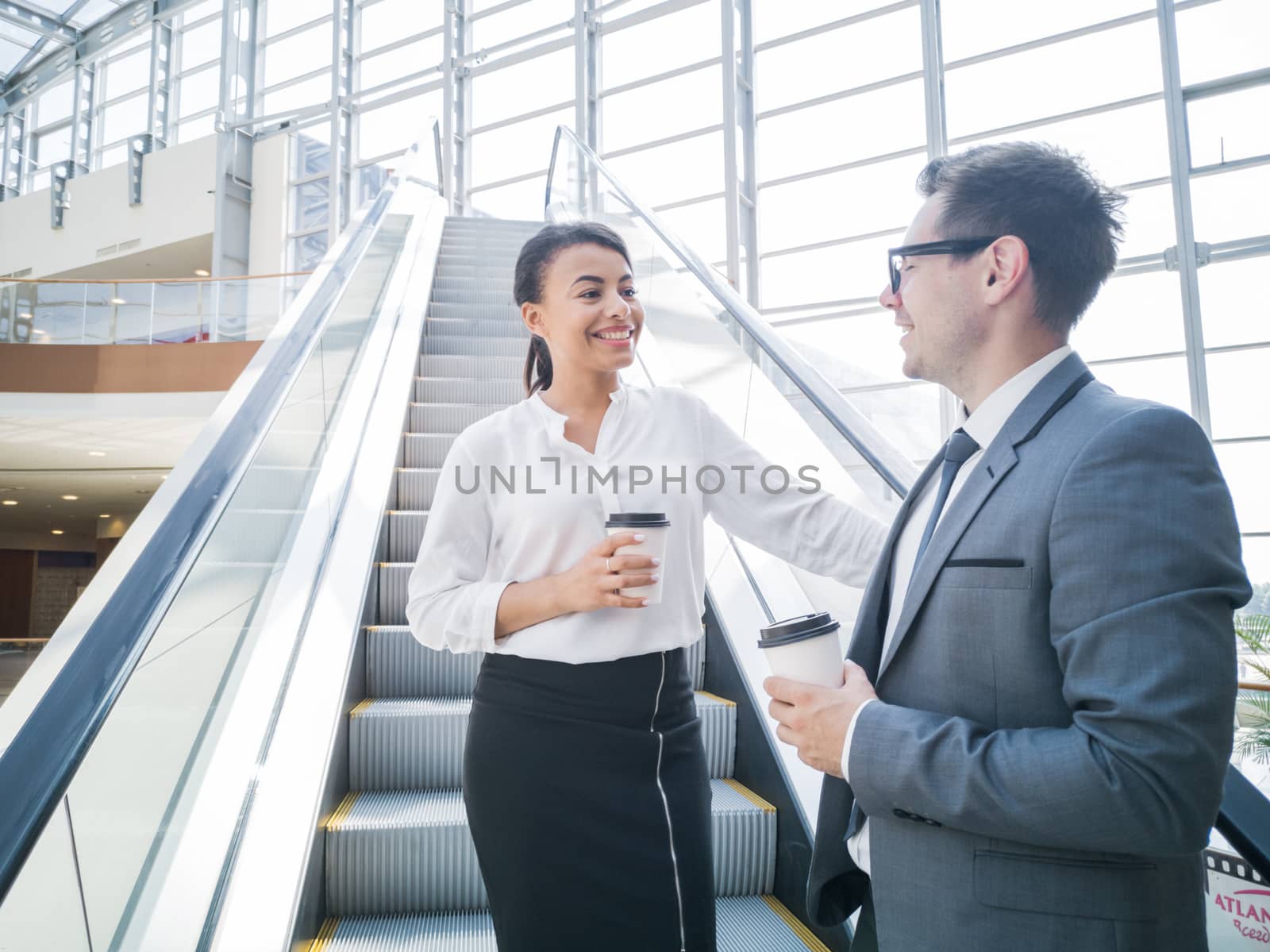 Businessman and businesswoman standing together on escalator and talk about business holding coffee in hand