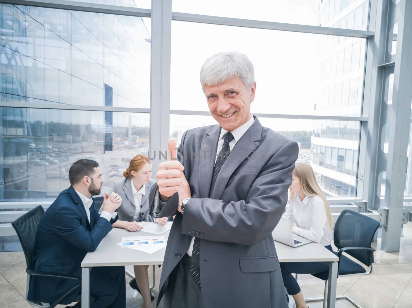 Confident mature businessman showing thumb up and smiling , team of his colleagues sitting at the table in the background