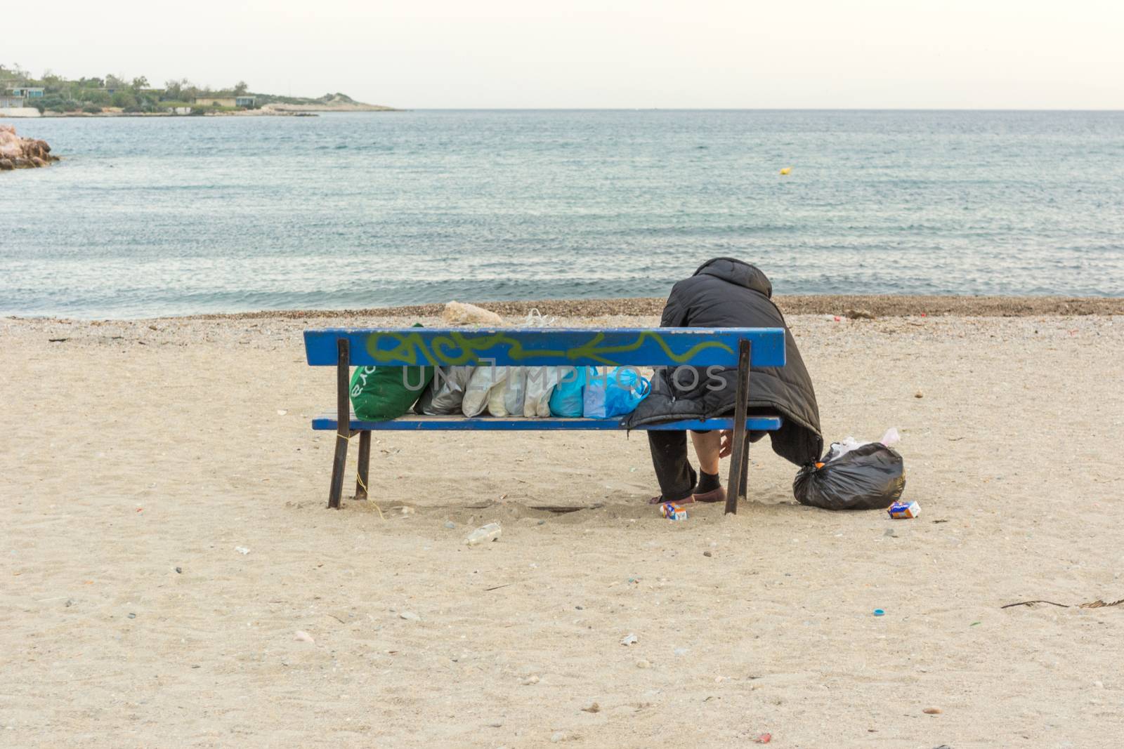 Homeless woman is sitting alone on a bench at the seashore with her stuff in bags, without hope, seashore, sandy beach