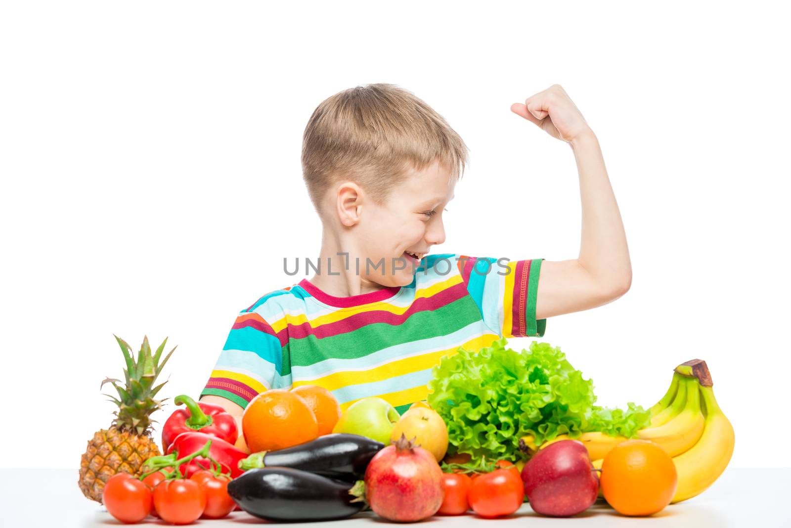 Strong boy shows biceps at the table with a pile of fresh vegetables and fruits isolated in the studio