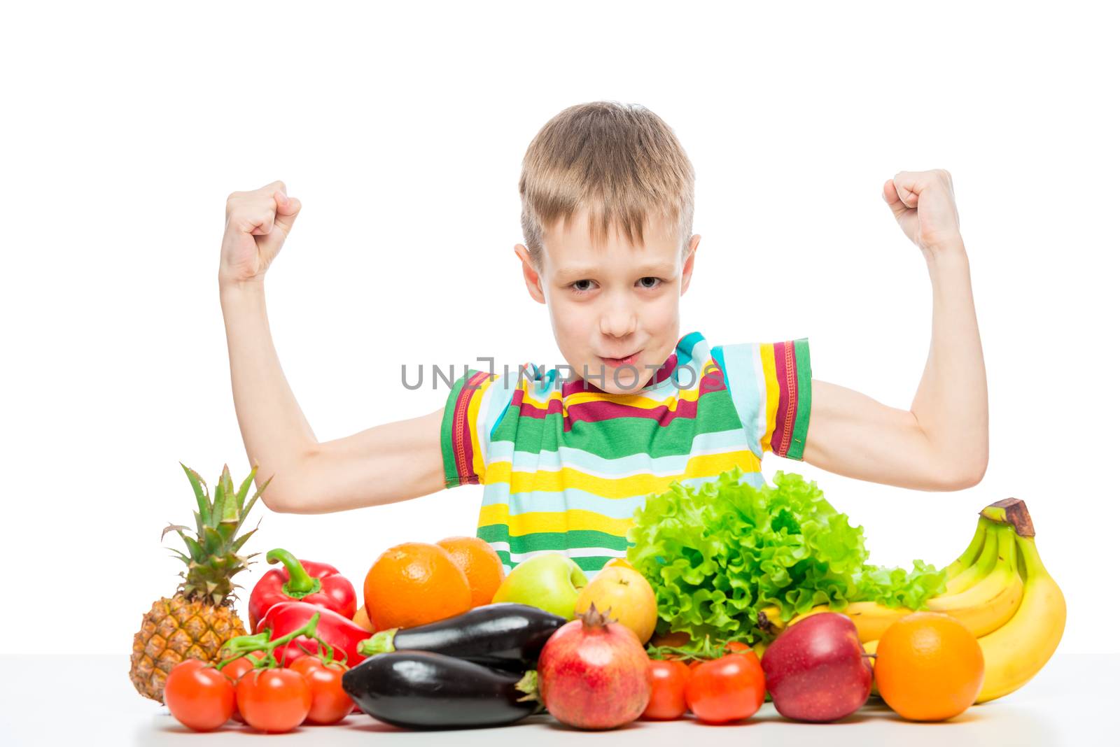 small strongman shows biceps at the table with a pile of fresh vegetables and fruits isolated in the studio