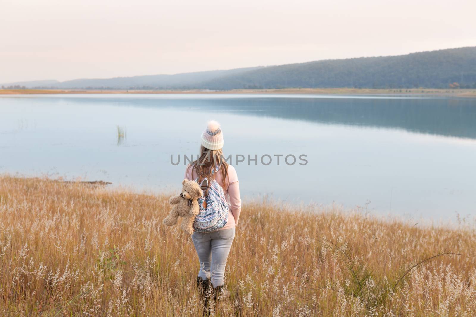 Woman wandering in long soft feather reed grass with wheat like spikes that gently sway in the breeze near a tranquil lake with pink hues at sunset. Country life