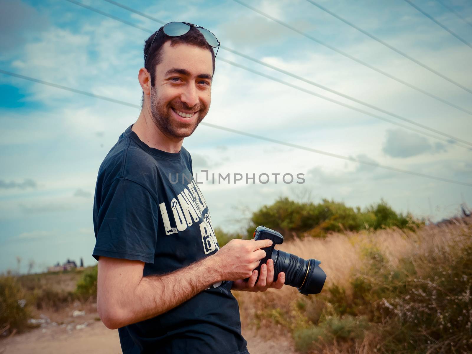 Happy young caucasian man photographer holding a digital camera in his hand and smiling to the camera outdoor in the field.