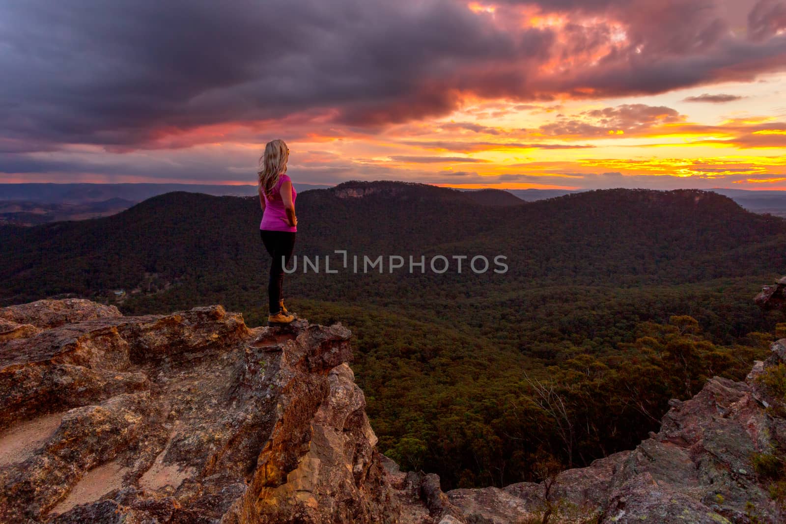 Storm clouds hover over Blue Mountains and valleys as the sun sets.  A woman stands on the rocky precipice taking in the magnificent views