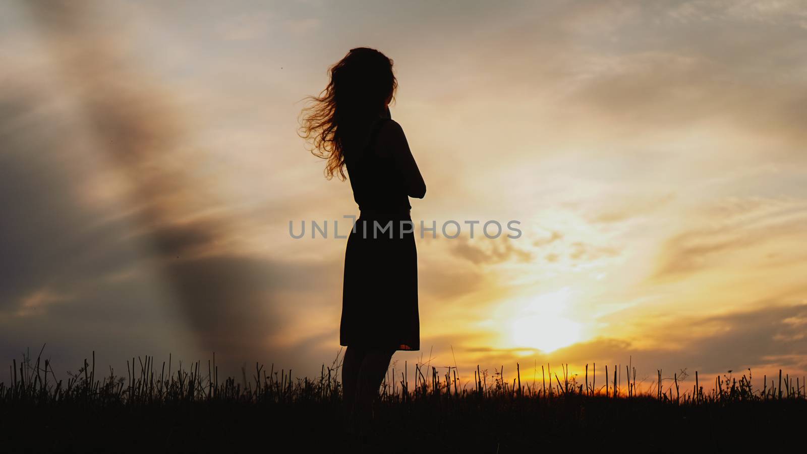 Silhouette of a young woman standing in dry grass field on bright sunset