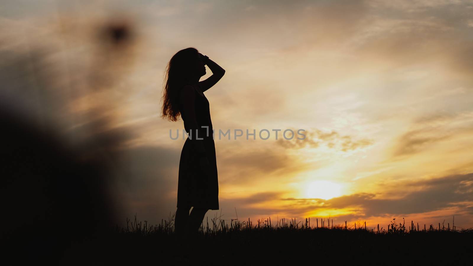 Silhouette of a young woman standing in dry grass field on bright sunset
