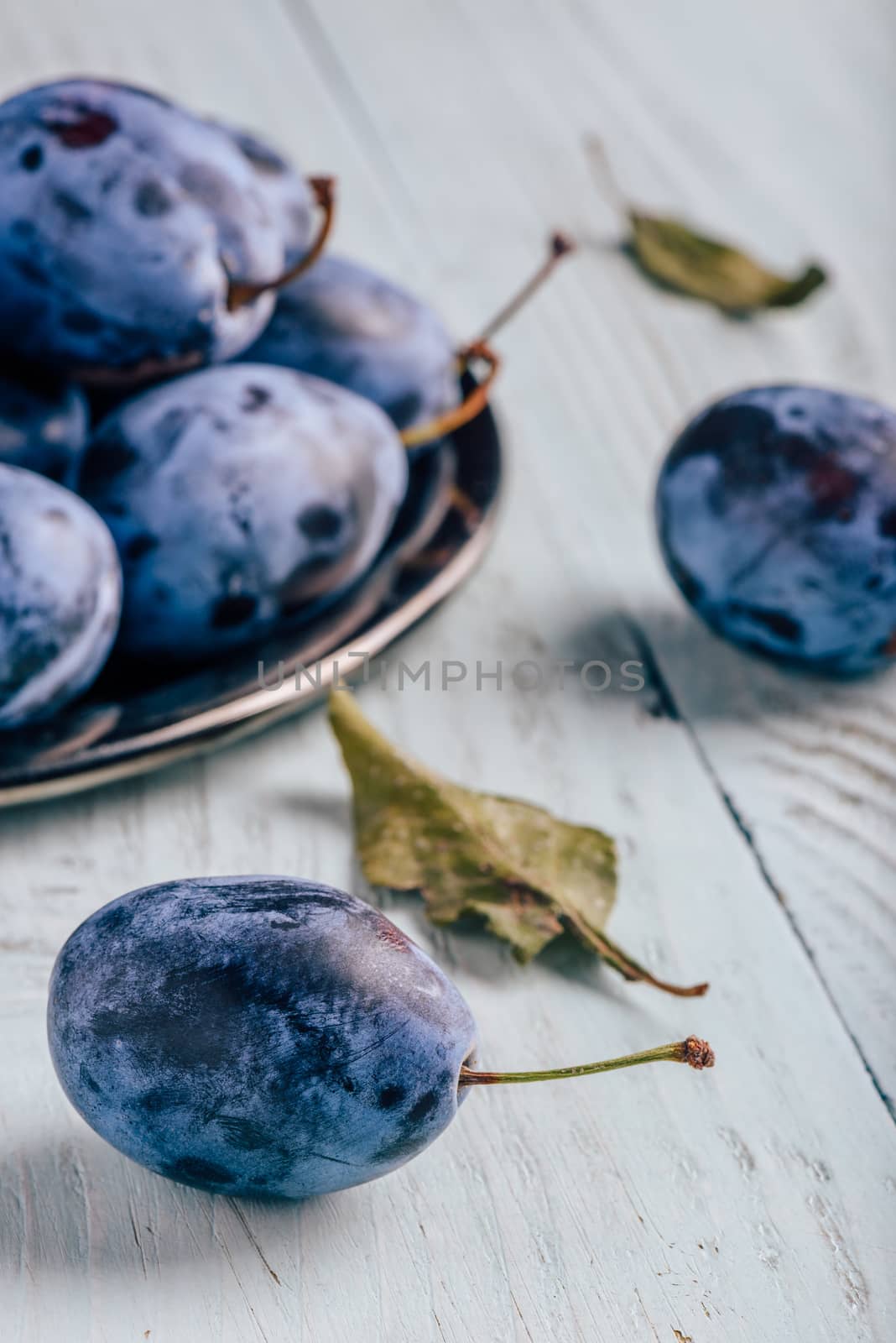 Ripe plums on metal plate and light wooden surface with leaves