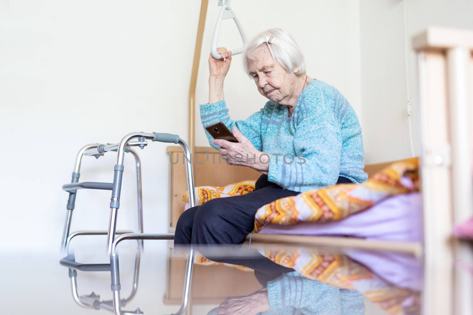 Elderly 96 years old woman reading phone message while sitting on medical bed supporting her by holder. Senior woman using wireless internet connection on smart phone.