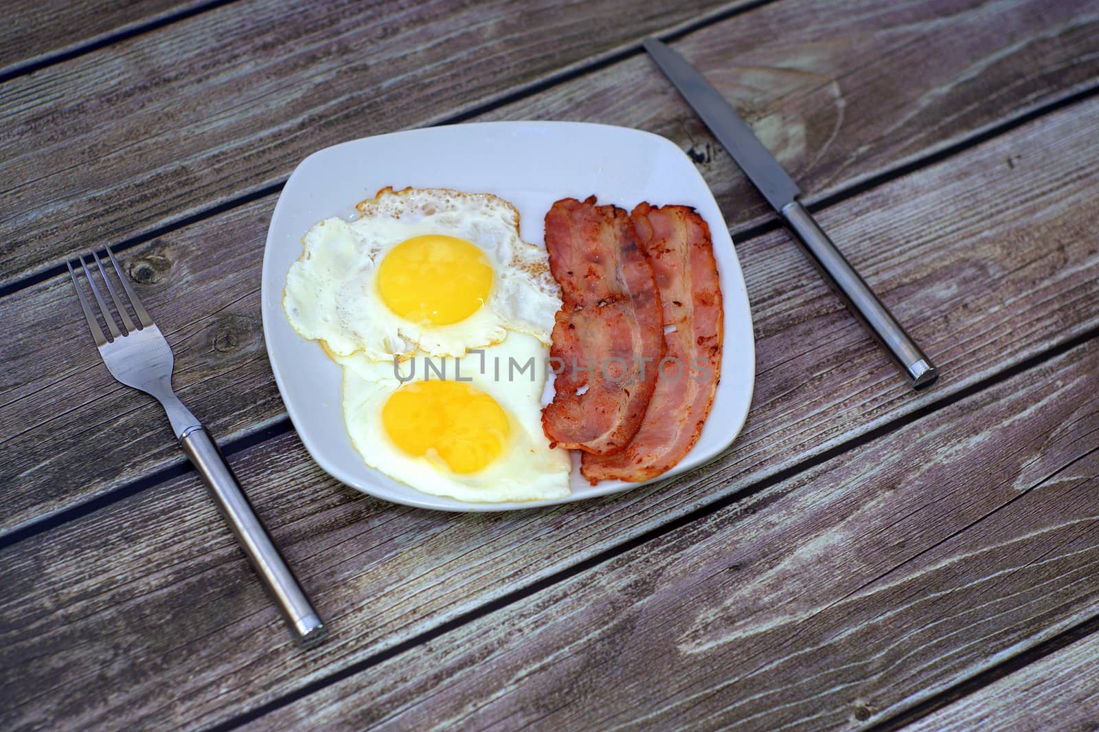 Traditional breakfast, plate with fried eggs and bacon and cutlery next to the table.