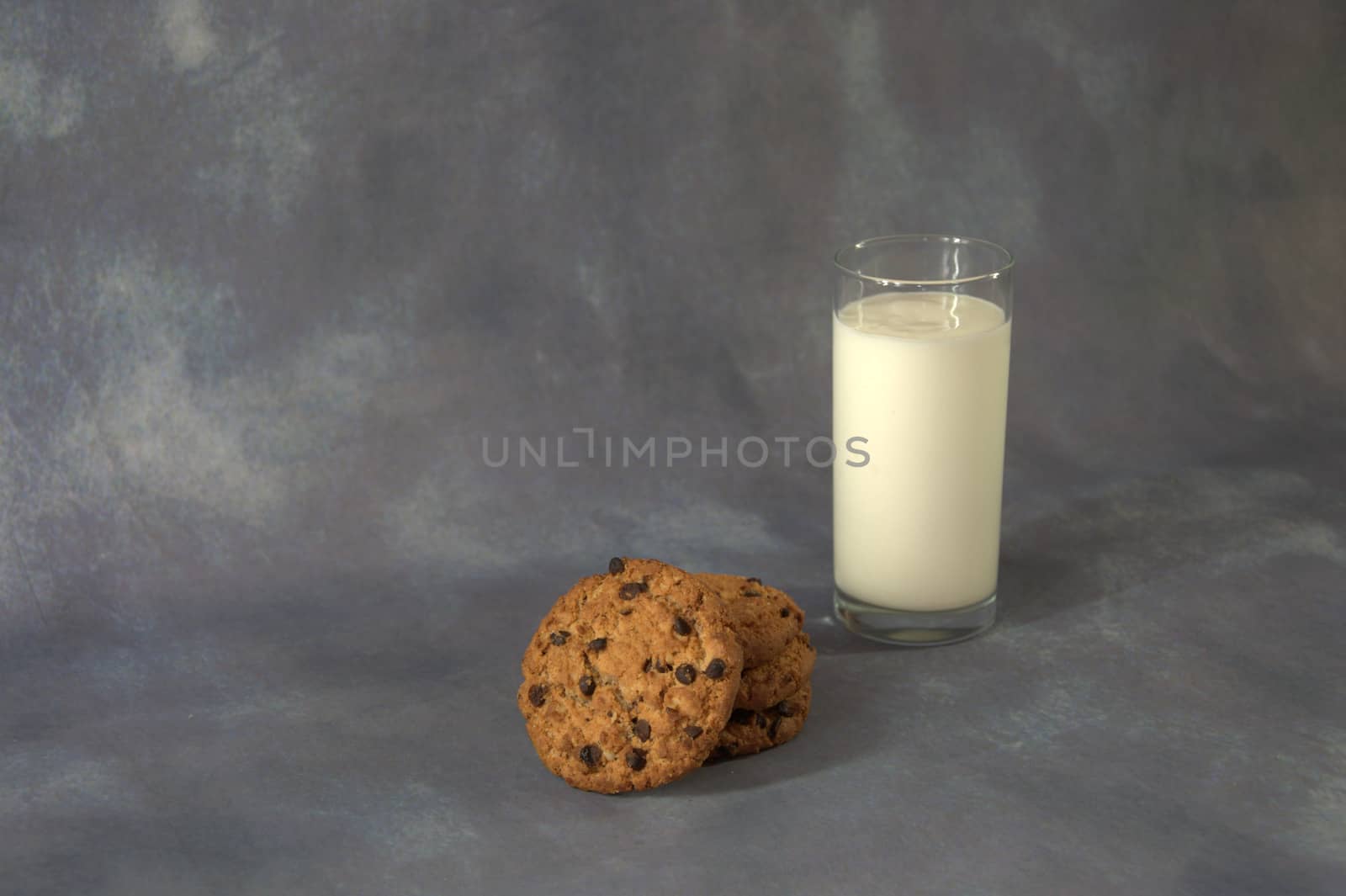 Full glass cup of milk and a few oatmeal cookies with chocolate on a gray abstract background.