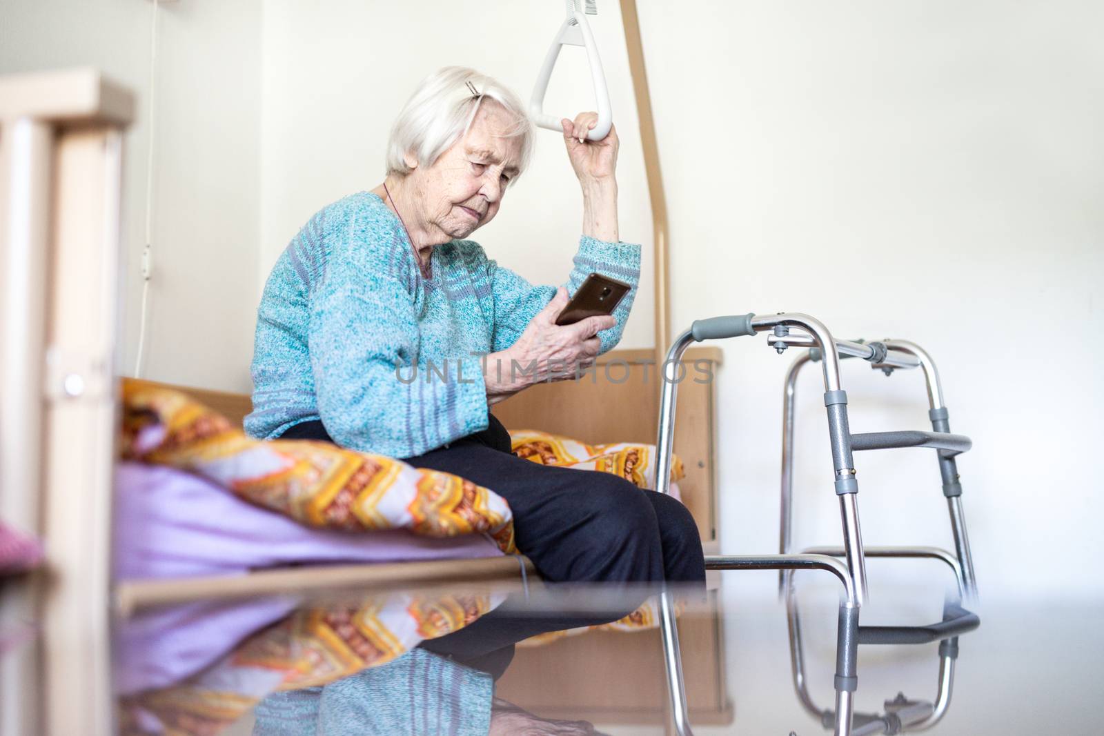 Elderly 96 years old woman reading phone message while sitting on medical bed supporting her by holder. Senior woman using wireless internet connection on smart phone.