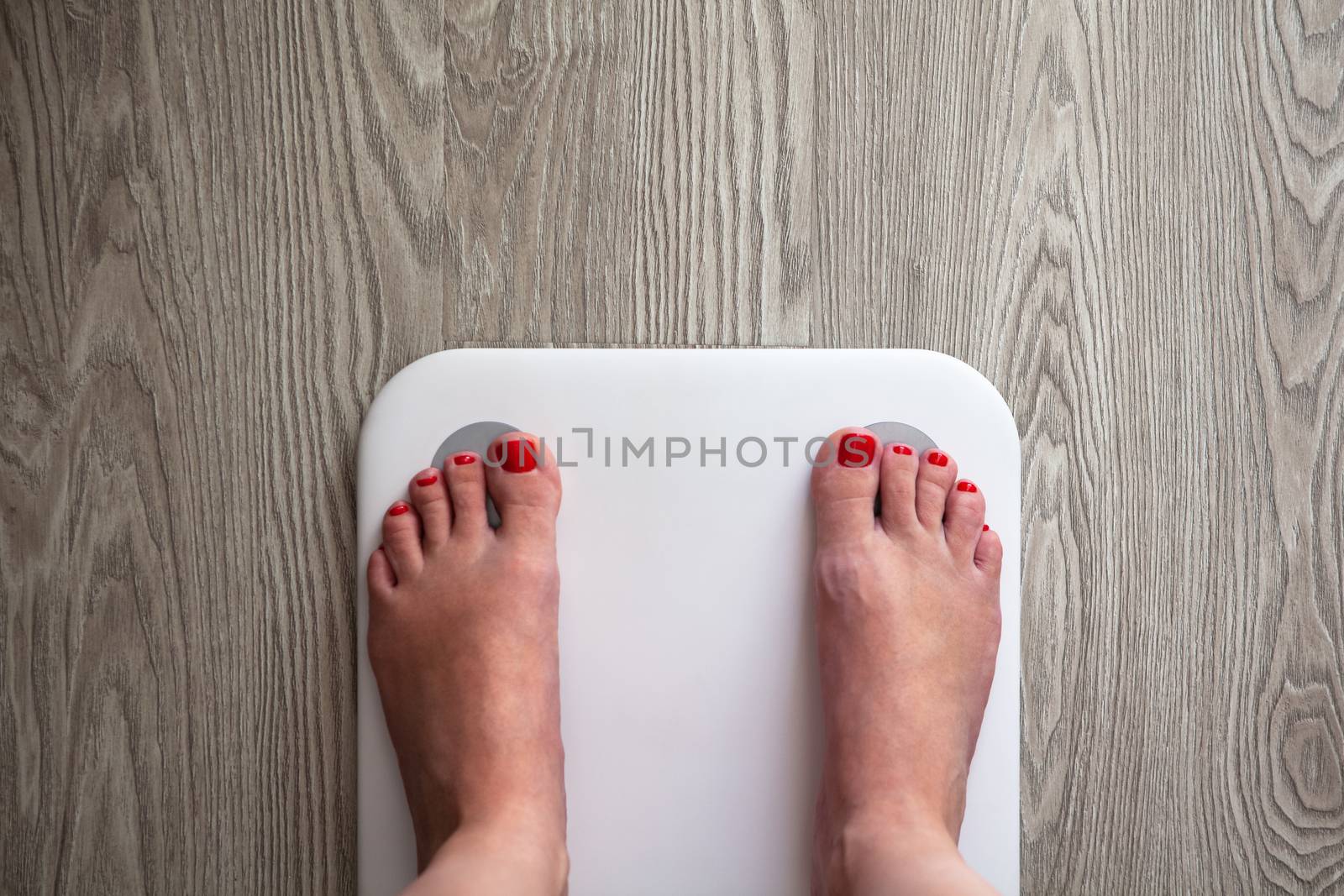 Woman stands on white modern electronic sensor scales. Only feet are visible. Scales stand on gray wooden floor. Copy space. Healthy lifestyle, diet, weight loss concept. Top view.