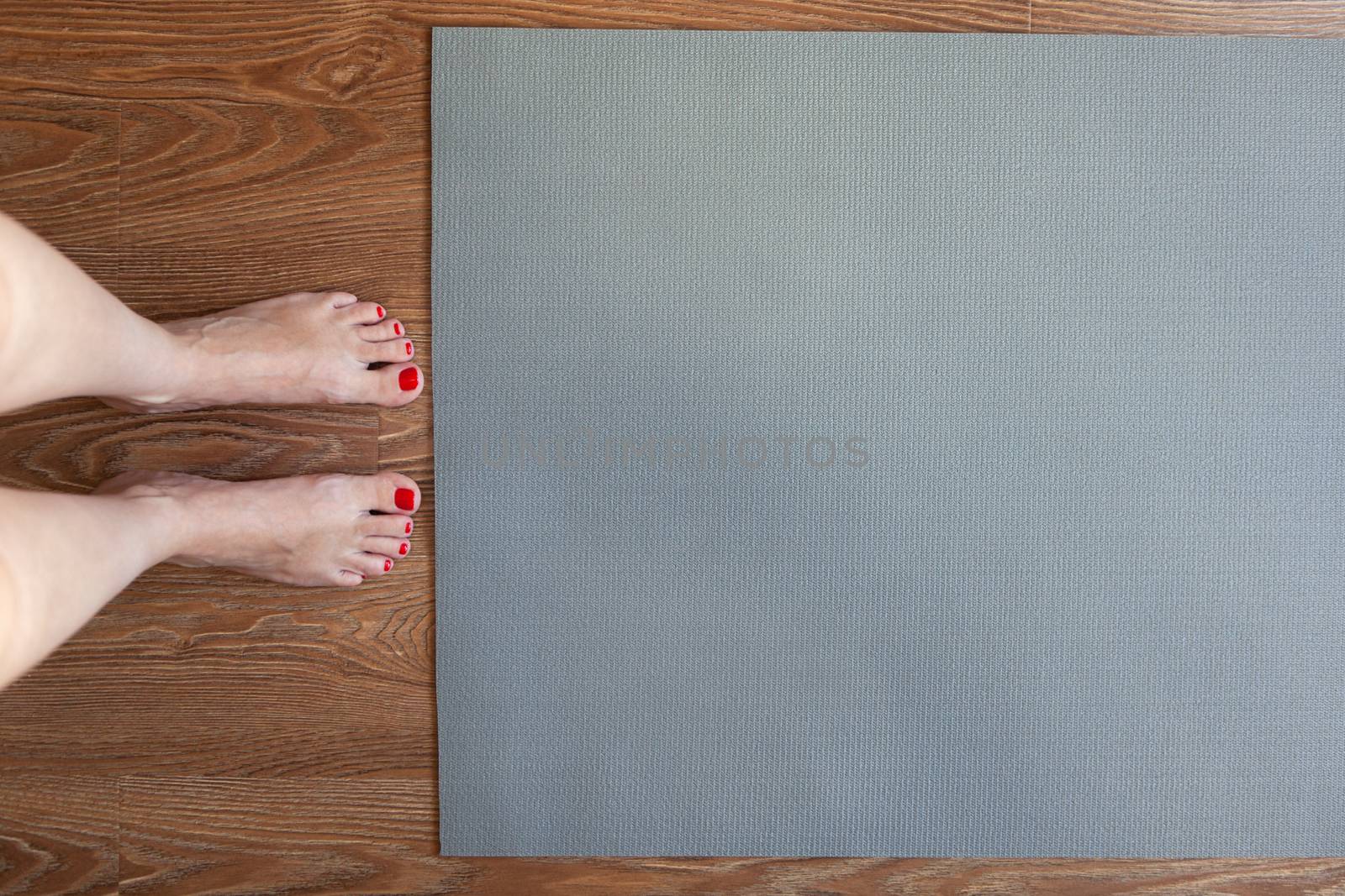 Woman is standing barefoot on floor in front of gymnastic mat, she is going to do morning exercise complex. Only legs are visible. Healthy lifestyle, weight loss concept. Horizontal. Copy space.