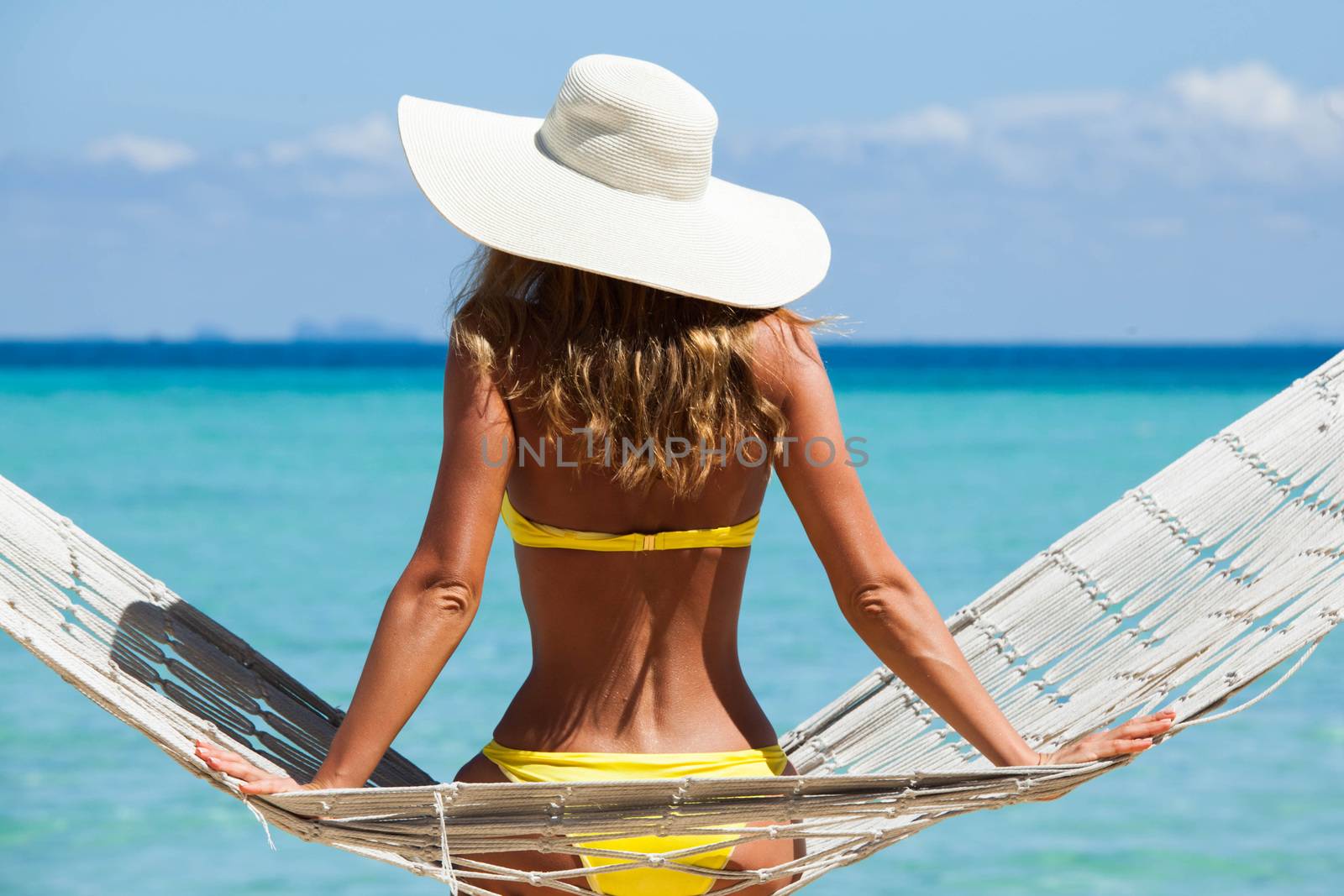 Rear view of a woman in bikini with a stylish hat sitting on a hammock on a beach by the sea