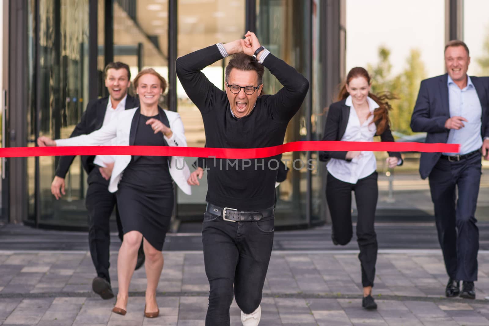 Group of happy business people running from office building crossing red ribbon finish line